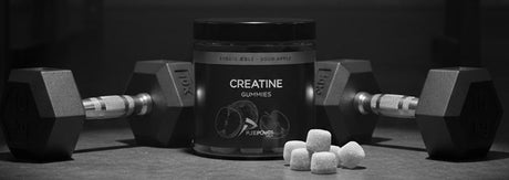 Sort/hvidt produktfoto af PurePower creatine gummies i åben beholder med flere vingummier foran, placeret mellem to håndvægte på træningsgulv — visuelt signalerer kreatin vingummi som et praktisk supplement til styrketræning og nem daglig dosering.