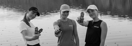 Tre atleter i træningstøj ved vandet holder energy gels frem mod kameraet — visuelt symbol på energiplan og gel‑brug til halvmarathon, timing og fuelingstrategi under løb.