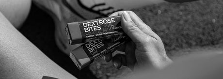 Sort-hvidt close-up af en hånd, der holder to Dextrose Bites energiprodukter med løbesko og træningsmiljø i baggrunden — illustrerer en hurtig, kulhydratrig snack til race-morning (top-up 30–60 min før marathon).