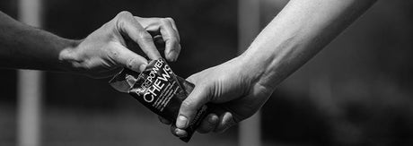 Sort-hvidt close-up af to hænder, der giver et PurePower Chews-energiprodukt videre, som illustrerer energiindtag under træning og valget mellem chews med og uden koffein.